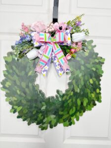 A faux boxwood wreath door hanger decorated with pastel florals and a colorful multi-ribbon bow. The greenery is shaped into a classic wreath silhouette, topped with pink, purple, and white florals.