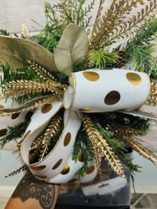 A gold and white polka-dotted ribbon bow with greenery, magnolia leaves, and golden ferns attached to a white candlestick. The foam is hidden beneath the full arrangement.