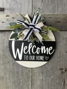 A round wooden door hanger with a 'Welcome to Our Home' design in black and white. The top left features a decorative bow made of layered ribbons in black, white, and natural burlap tones, accented with lace trim. Behind the bow, lush greenery, including ferns and eucalyptus, extends outward for added texture. The sign hangs against a rustic wooden background, creating a charming farmhouse-style entrance piece.
