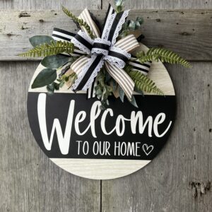 A round wooden door hanger with a 'Welcome to Our Home' design in black and white. The top left features a decorative bow made of layered ribbons in black, white, and natural burlap tones, accented with lace trim. Behind the bow, lush greenery, including ferns and eucalyptus, extends outward for added texture. The sign hangs against a rustic wooden background, creating a charming farmhouse-style entrance piece.
