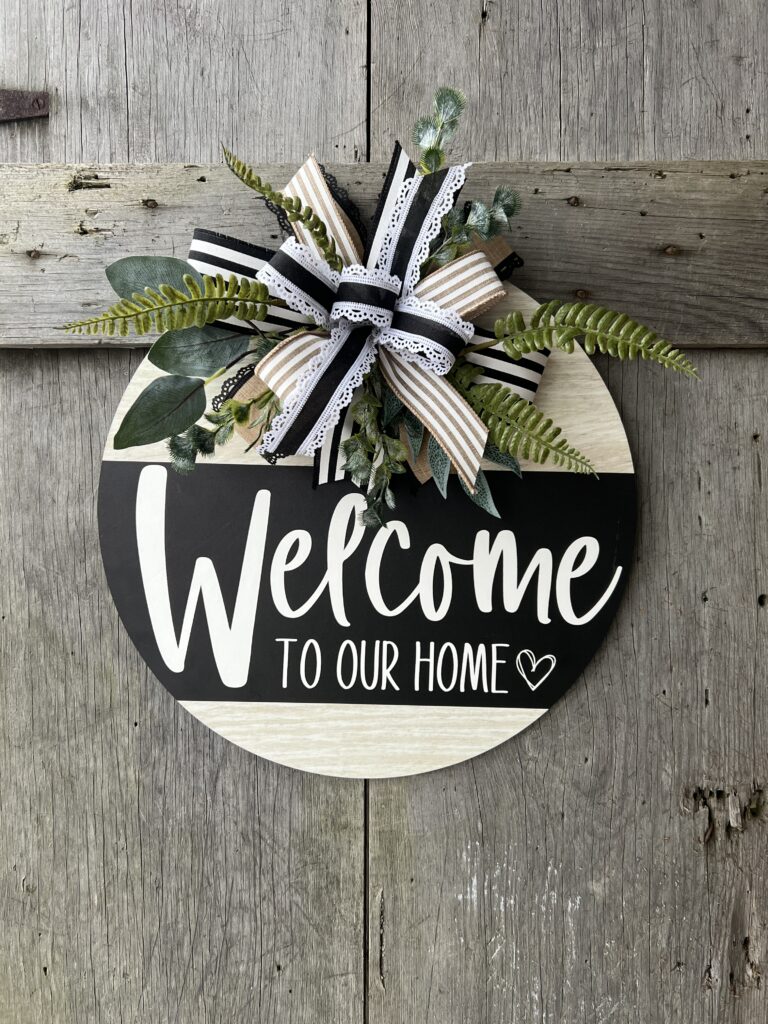 A round wooden door hanger with a 'Welcome to Our Home' design in black and white. The top left features a decorative bow made of layered ribbons in black, white, and natural burlap tones, accented with lace trim. Behind the bow, lush greenery, including ferns and eucalyptus, extends outward for added texture. The sign hangs against a rustic wooden background, creating a charming farmhouse-style entrance piece.