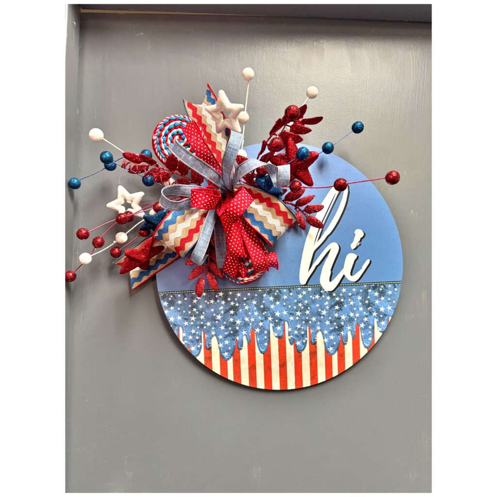 Full shot of a patriotic summer door hanger on a gray background, decorated with red, white, and blue elements, glittered floral picks, and a festive bow above the word “hi.”