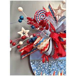 Close-up view of a red, white, and blue summer-themed door hanger, highlighting the layered patriotic bow with denim and ric rac ribbon, surrounded by glittered star picks and round berry sprays.